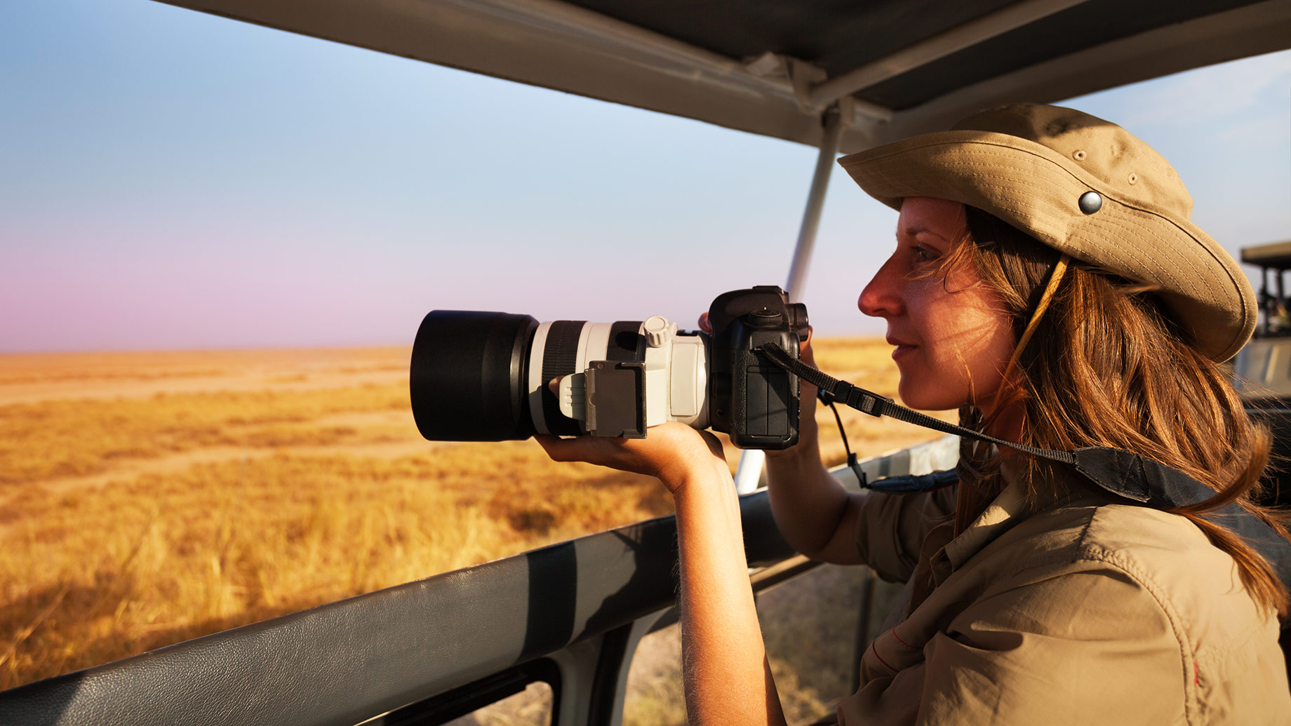 Woman holding a professional telephoto camera aboard an open safari vehicle overlooking the sunlit African savannah.