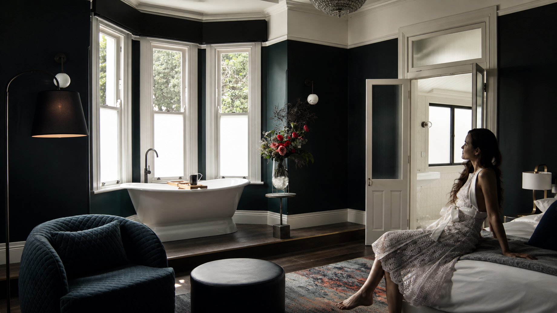Woman relaxing beside a freestanding bathtub in an elegant dark-toned bathroom with bay windows, warm lighting, and refined furnishings.