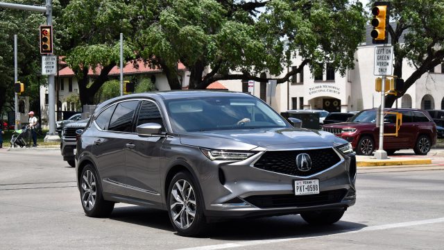 Grey Acura RDX SUV travelling through a Houston intersection beneath traffic signals on a sunny day.