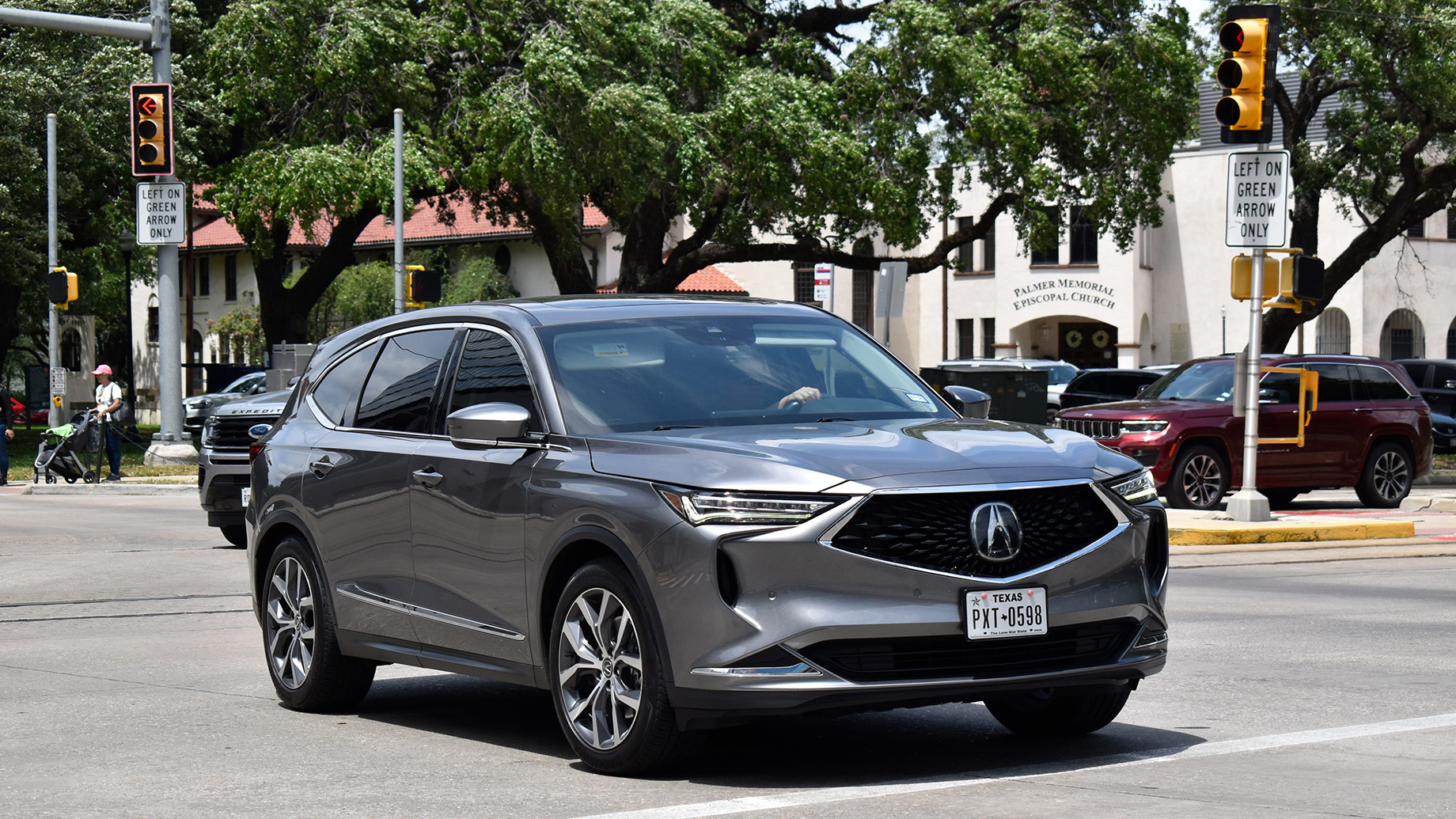 Grey Acura RDX SUV travelling through a Houston intersection beneath traffic signals on a sunny day.