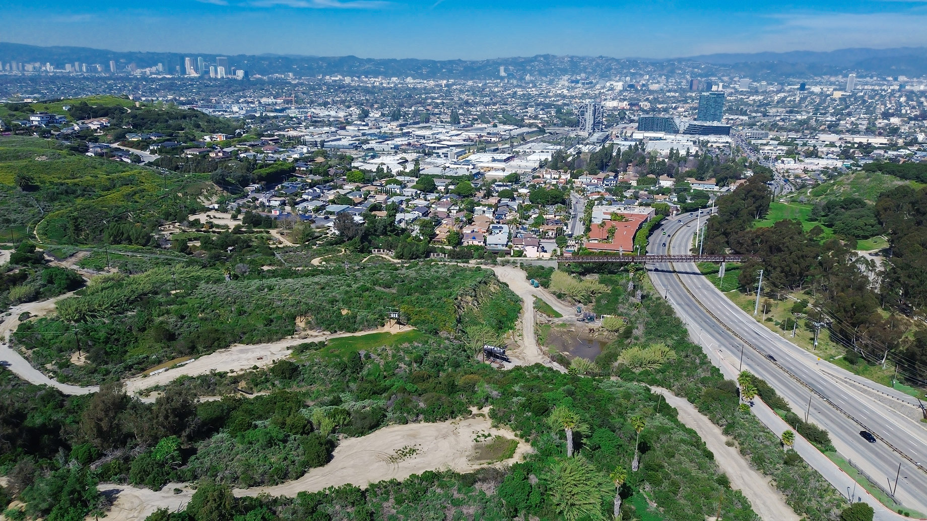 Aerial view from Kenneth Hahn State Recreation Area toward Culver City and the Los Angeles skyline with green hills and winding trails.