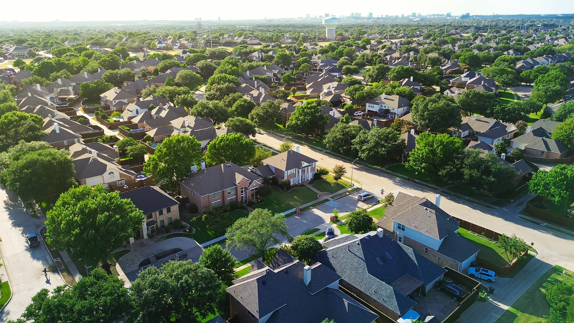 Aerial view of tree-lined suburban homes in West Plano, Texas, with a water tower and the distant Dallas skyline.