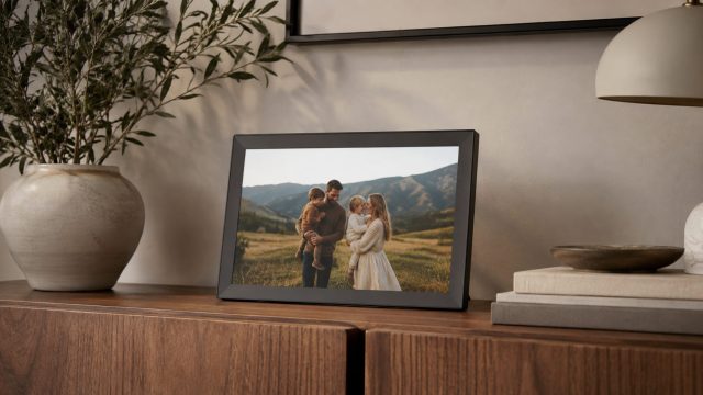 Matte electronic frame displaying a family memory on a walnut console with ceramic decor, books, and warm minimalist styling.