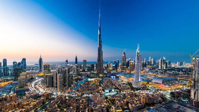 Elevated view of Downtown Dubai at twilight with the Burj Khalifa rising above illuminated towers, residential districts, and surrounding city streets.