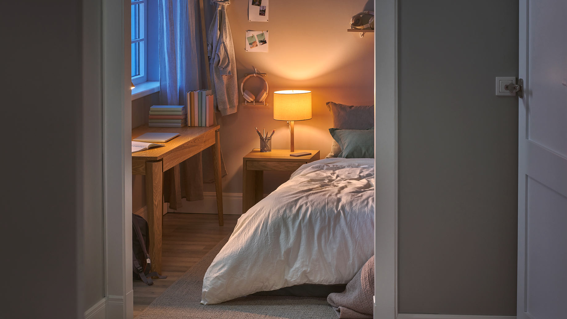 Cozy teenage bedroom with white bedding, a warm bedside lamp, wooden study desk, books, and soft evening light entering through the window.