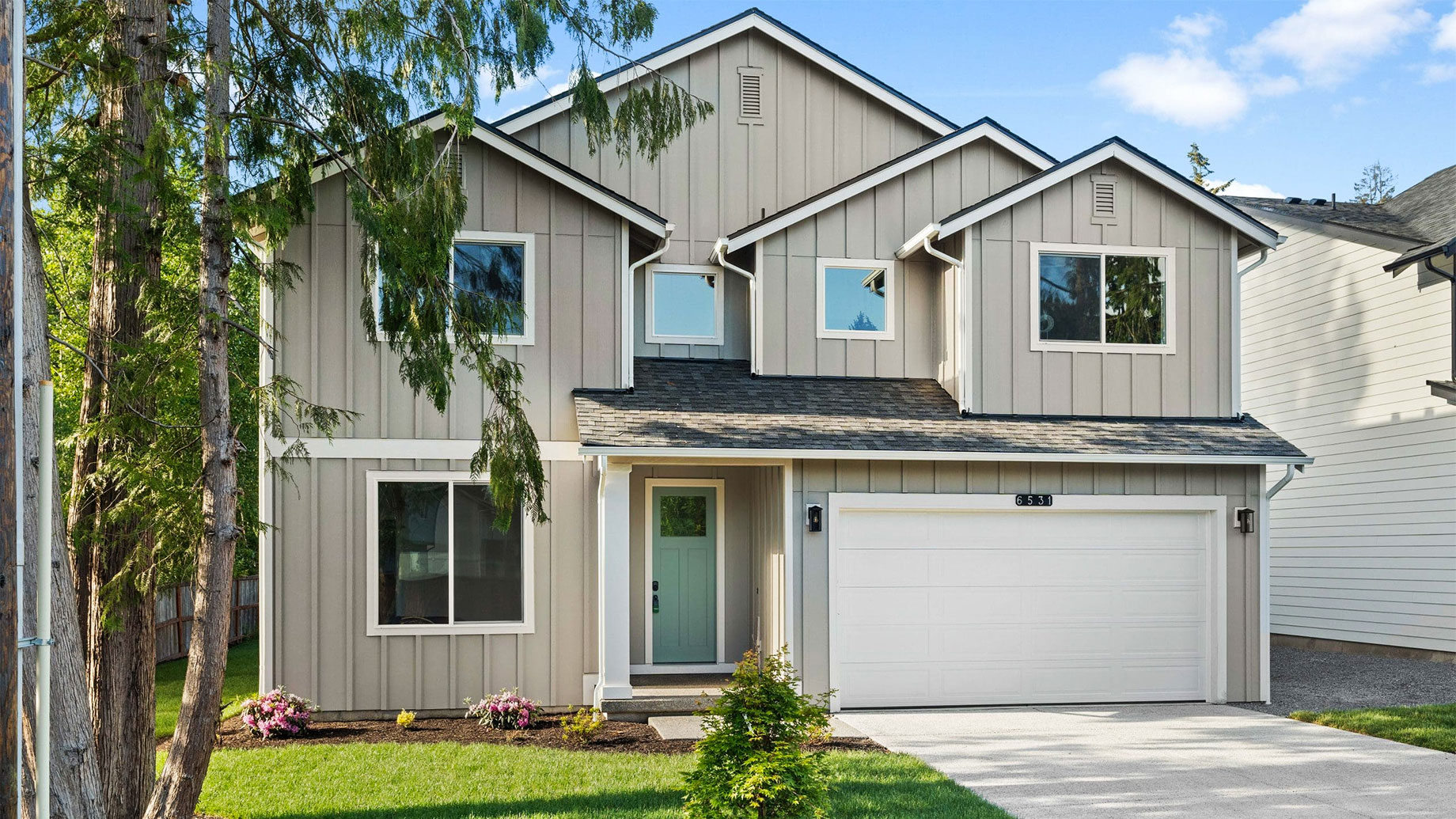 Two-story suburban home with gray siding, multiple gabled roofs, a sage-green front door, white garage door, and concrete driveway surrounded by evergreen trees.