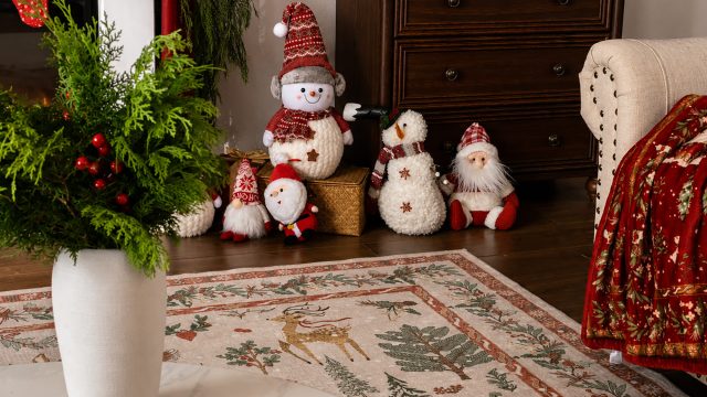 Cozy Christmas living room with a festive reindeer rug, evergreen greenery, snowmen, Santa decorations, red textiles, and warm wood furniture.