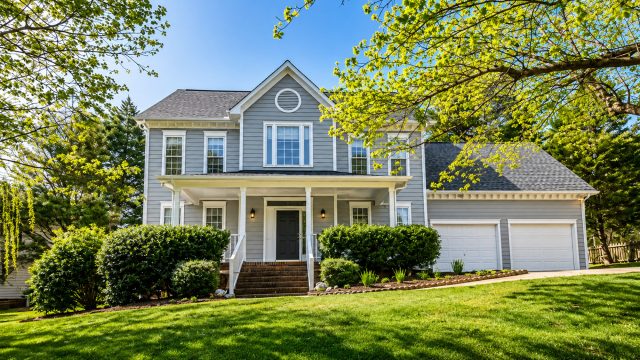 Two-story suburban rental property with grey siding, an attached garage, mature trees, and a landscaped front yard.