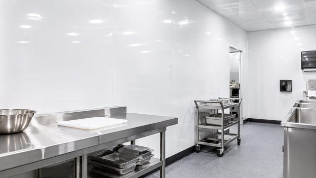 Commercial food preparation space with seamless white wall panels, stainless-steel work surfaces, sinks, and a mobile equipment cart.
