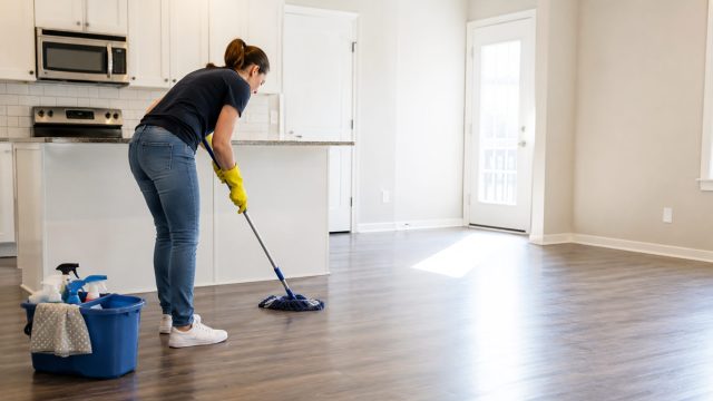Cleaner mopping the floor of an empty open-plan home with cleaning supplies arranged beside the kitchen.