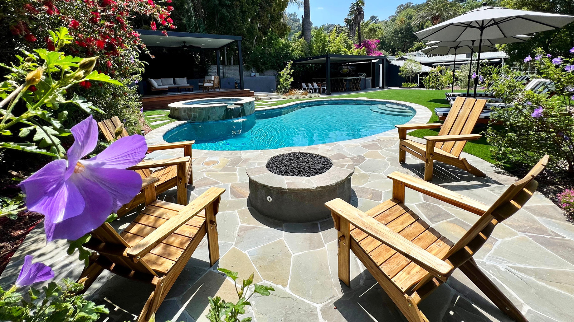 Flagstone patio surrounding a swimming pool with wooden lounge chairs, a circular fire pit, shaded seating, and colourful garden landscaping.