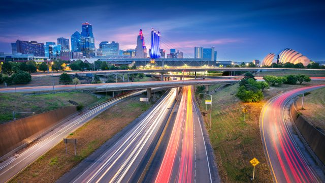 Kansas City skyline at dusk above intersecting highways with long-exposure traffic light trails.
