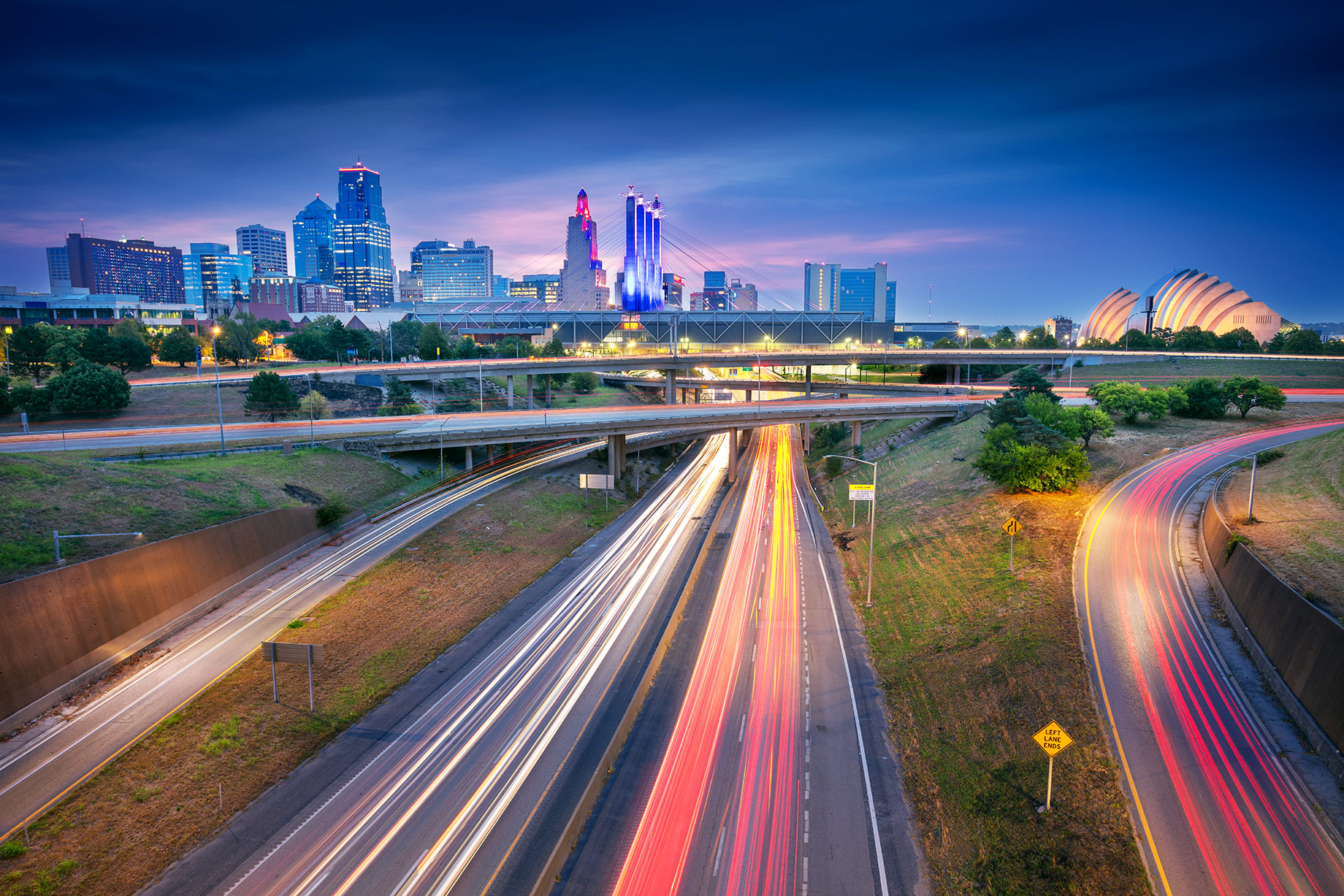 Kansas City skyline at dusk above intersecting highways with long-exposure traffic light trails.