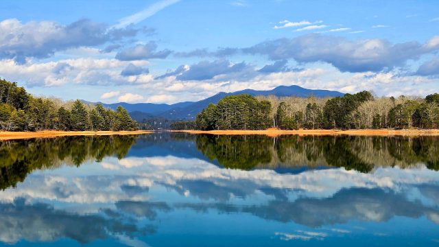 Calm waters of Lake Chatuge reflecting forested shoreline, Blue Ridge mountain peaks, and a partly cloudy sky in western North Carolina.