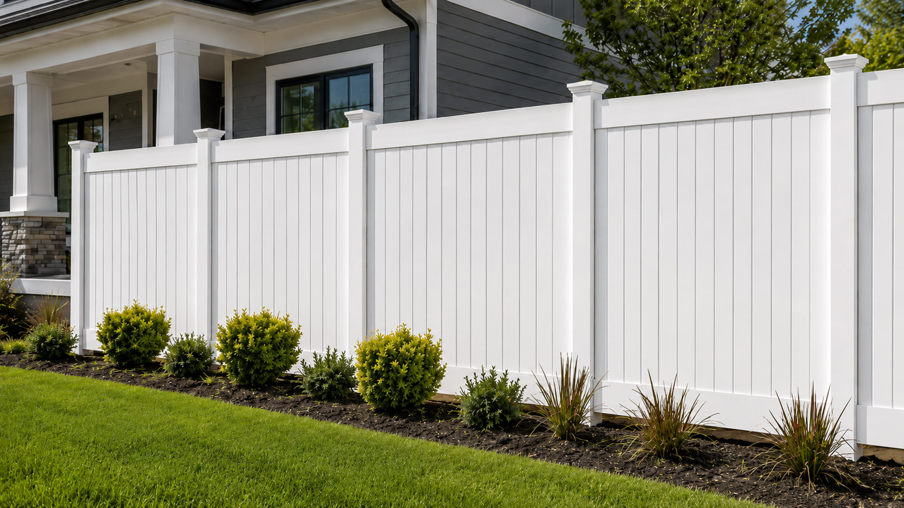 Clean white privacy fence with evenly spaced vertical panels, landscaped garden beds, green lawn, and a modern residential home in the background.