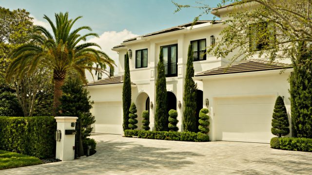 Luxury white mansion with a gated entrance, palm trees, manicured landscaping, and a spacious driveway.