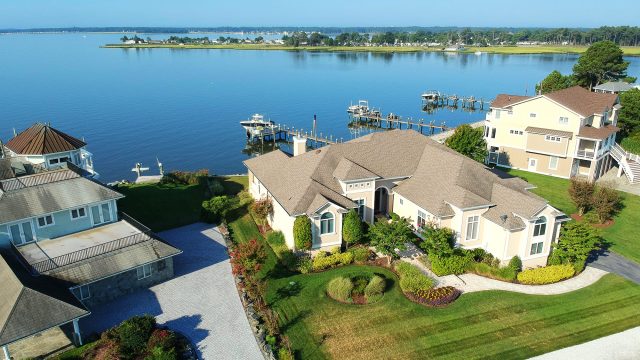 Aerial view of luxury waterfront homes with broad shingle roofs, landscaped grounds, private docks, and a calm coastal inlet.
