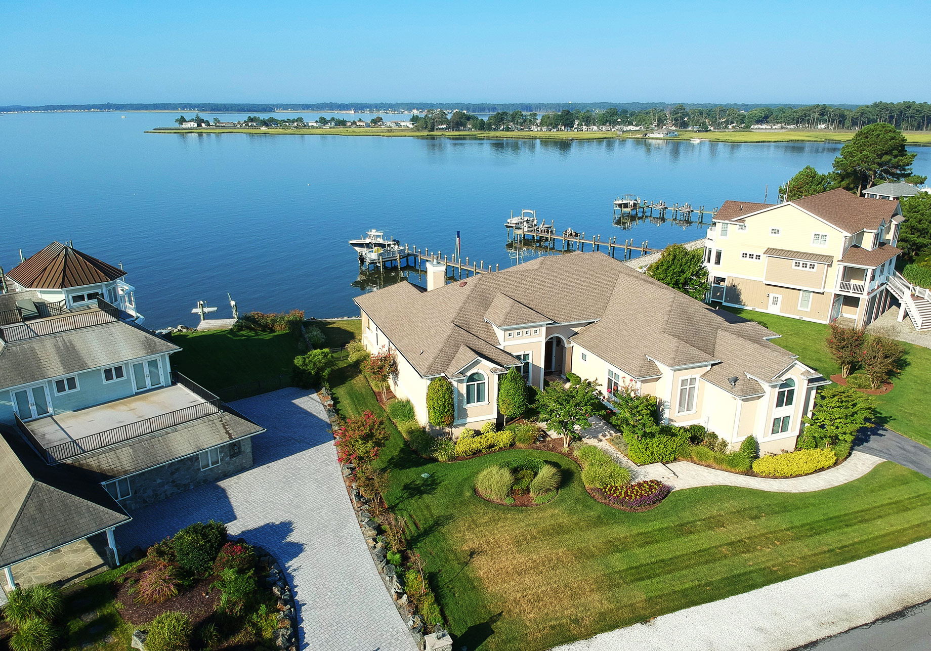 Aerial view of luxury waterfront homes with broad shingle roofs, landscaped grounds, private docks, and a calm coastal inlet.