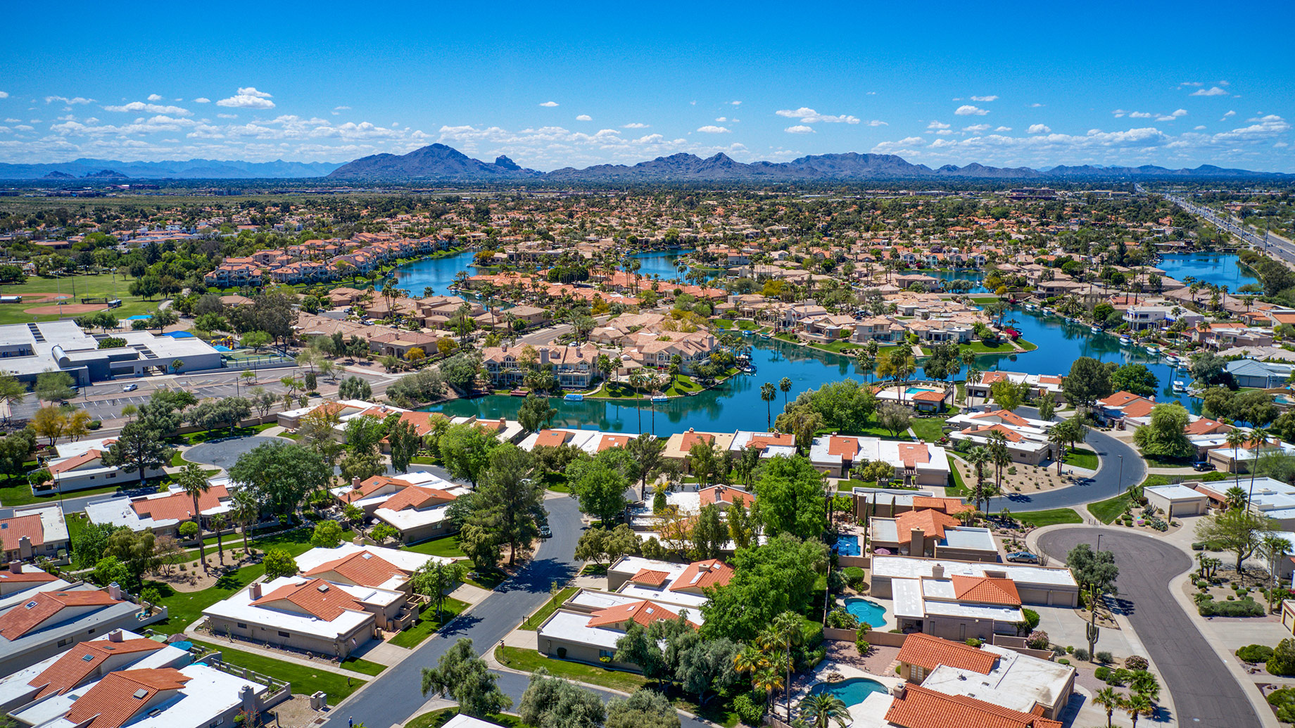 Aerial view of a North Scottsdale neighborhood with residential homes, community lakes, palm trees, and desert mountains under a clear blue sky.