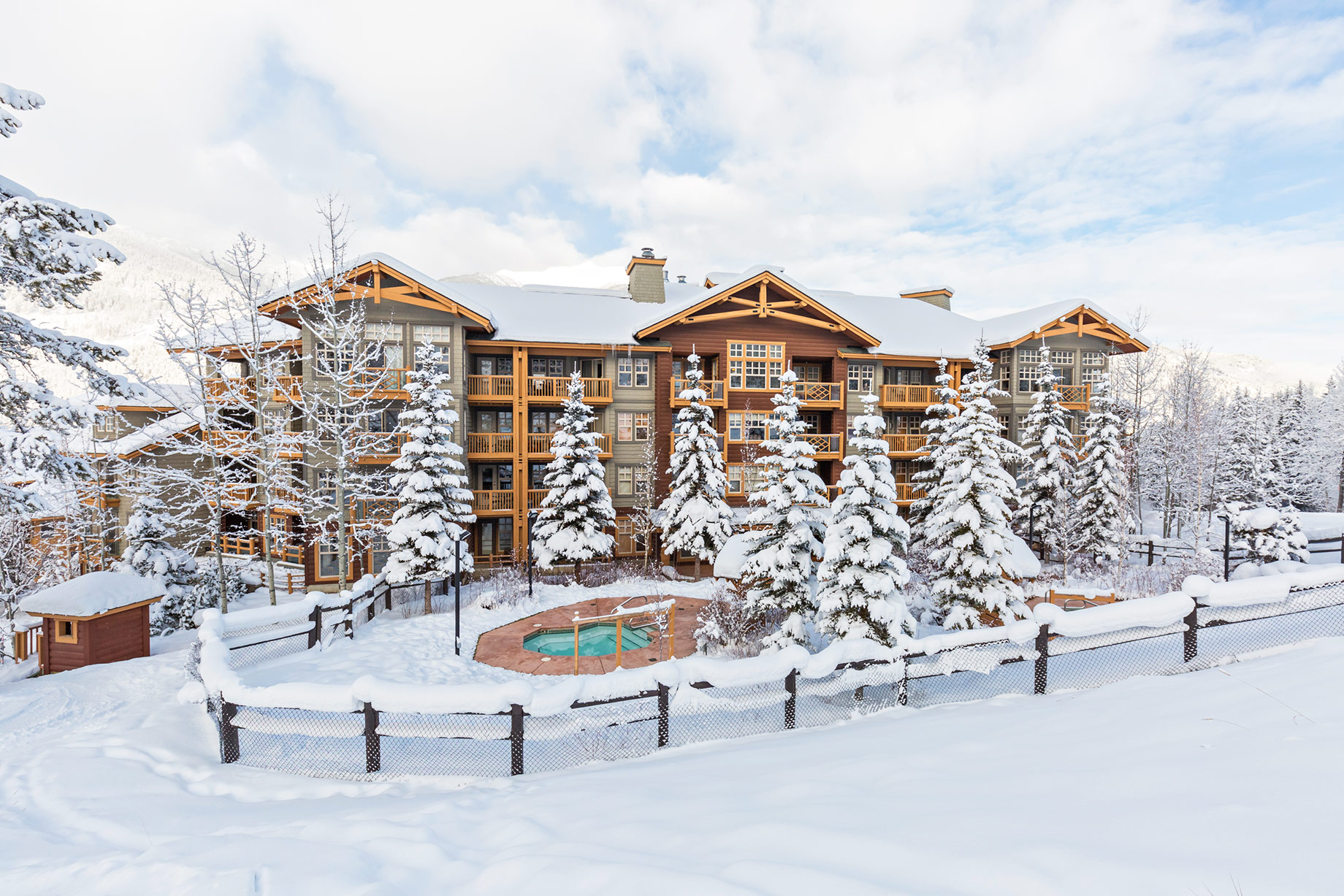 Multi-storey ski resort condominiums with balconies, snow-covered evergreens, and an outdoor hot tub.