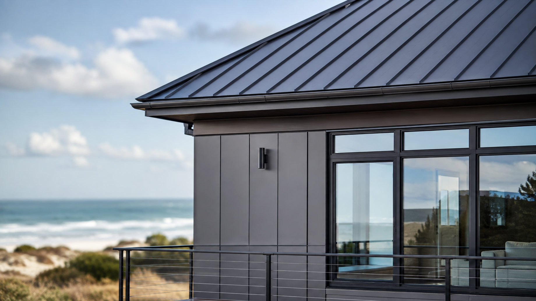 Close-up of a modern steel coastal home with standing-seam metal roofing, evenly spaced exterior panels, large windows, and an ocean backdrop.