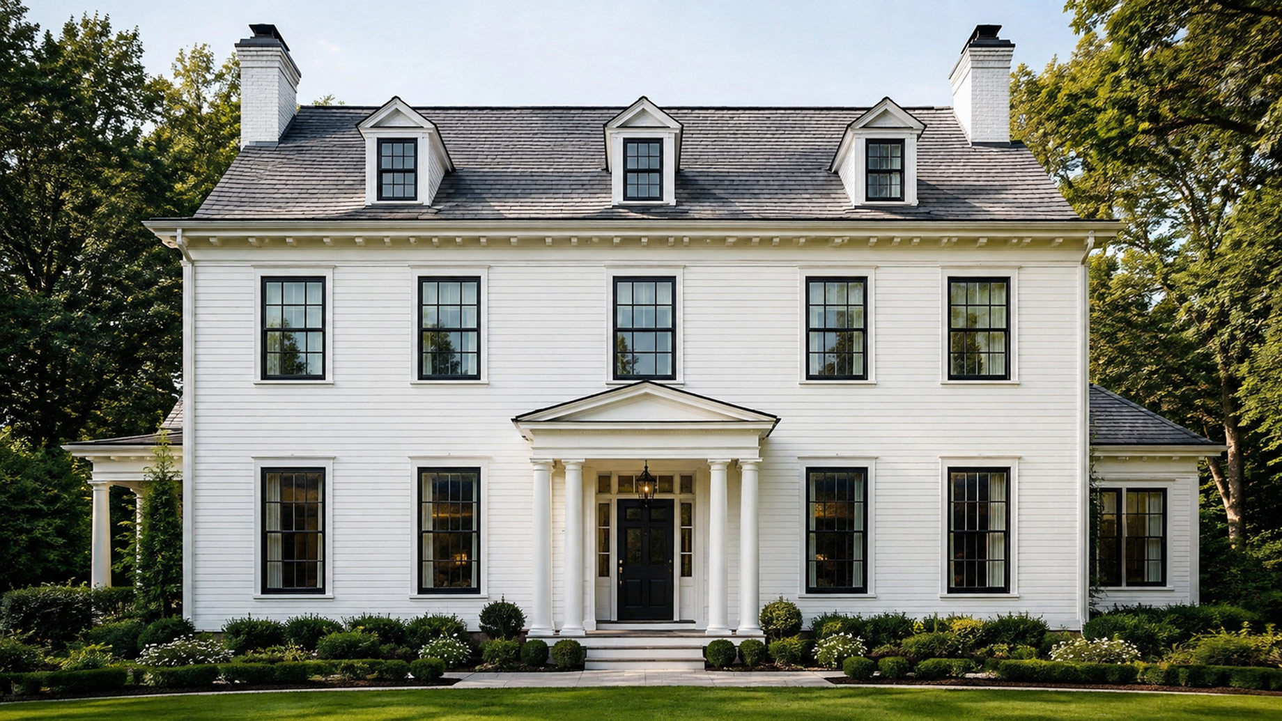 Symmetrical traditional white home with black-framed casement windows, dormers, classical entry columns, and formal landscaping.