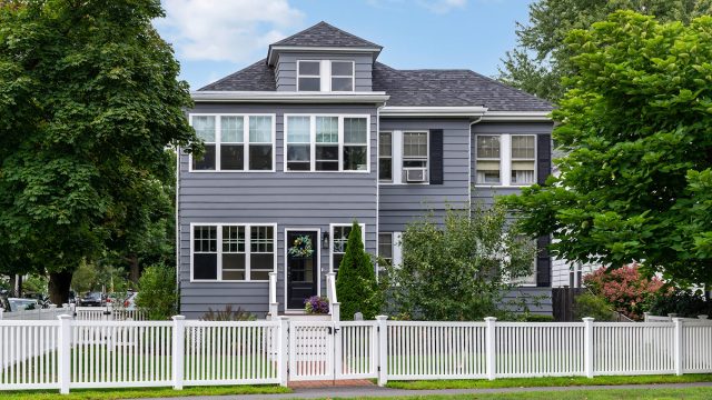 Traditional grey two-story home with white trim, mature trees, landscaped gardens, and a white picket fence in Watertown, Massachusetts.