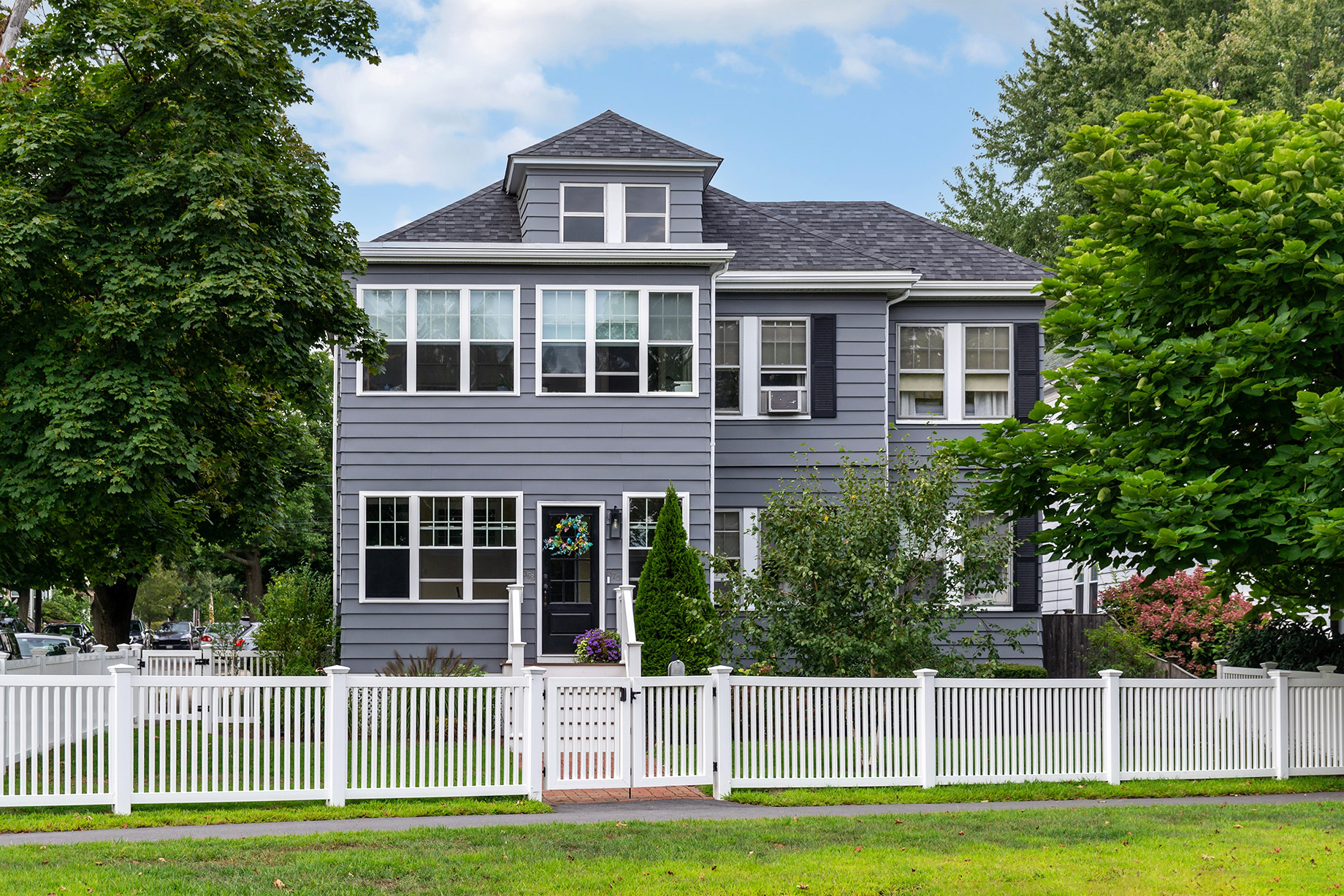 Traditional grey two-story home with white trim, mature trees, landscaped gardens, and a white picket fence in Watertown, Massachusetts.