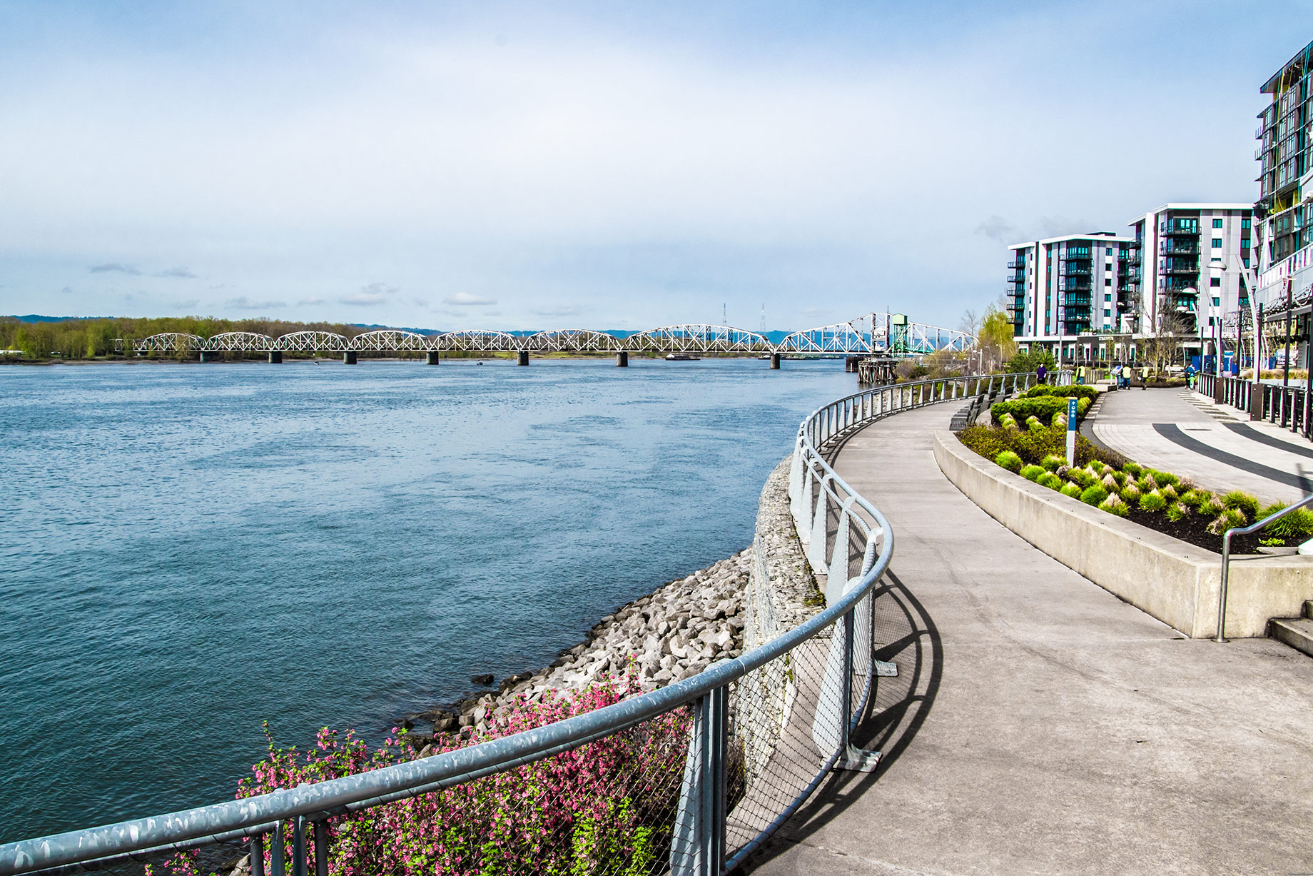 Curving riverwalk along the Columbia River in Vancouver, Washington, with landscaped gardens, modern waterfront buildings, and a bridge in the distance.