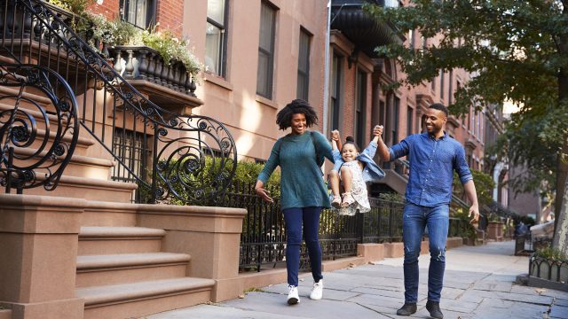 Family walking together along a tree-lined Brooklyn street bordered by historic brownstone homes.