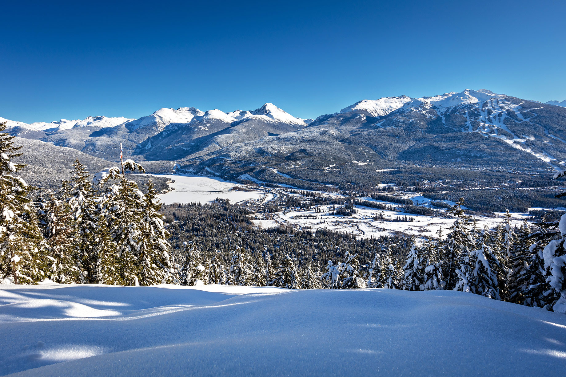 Snow-covered Whistler Valley with Blackcomb Mountain, Wedge Mountain, Mount Weart, and Arm Chair Glacier beneath a clear blue winter sky.