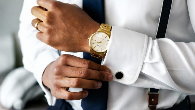 Young professional adjusting a crisp white shirt cuff while wearing a gold luxury watch, navy tie, suspenders, and formal business attire.