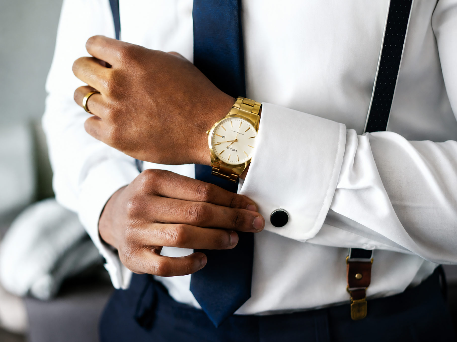 Young professional adjusting a crisp white shirt cuff while wearing a gold luxury watch, navy tie, suspenders, and formal business attire.