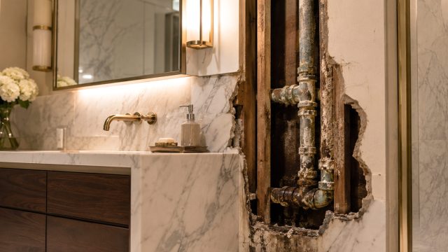 Corroded metal plumbing exposed behind marble wall finishes in an upscale bathroom with a custom vanity and brass fixtures.