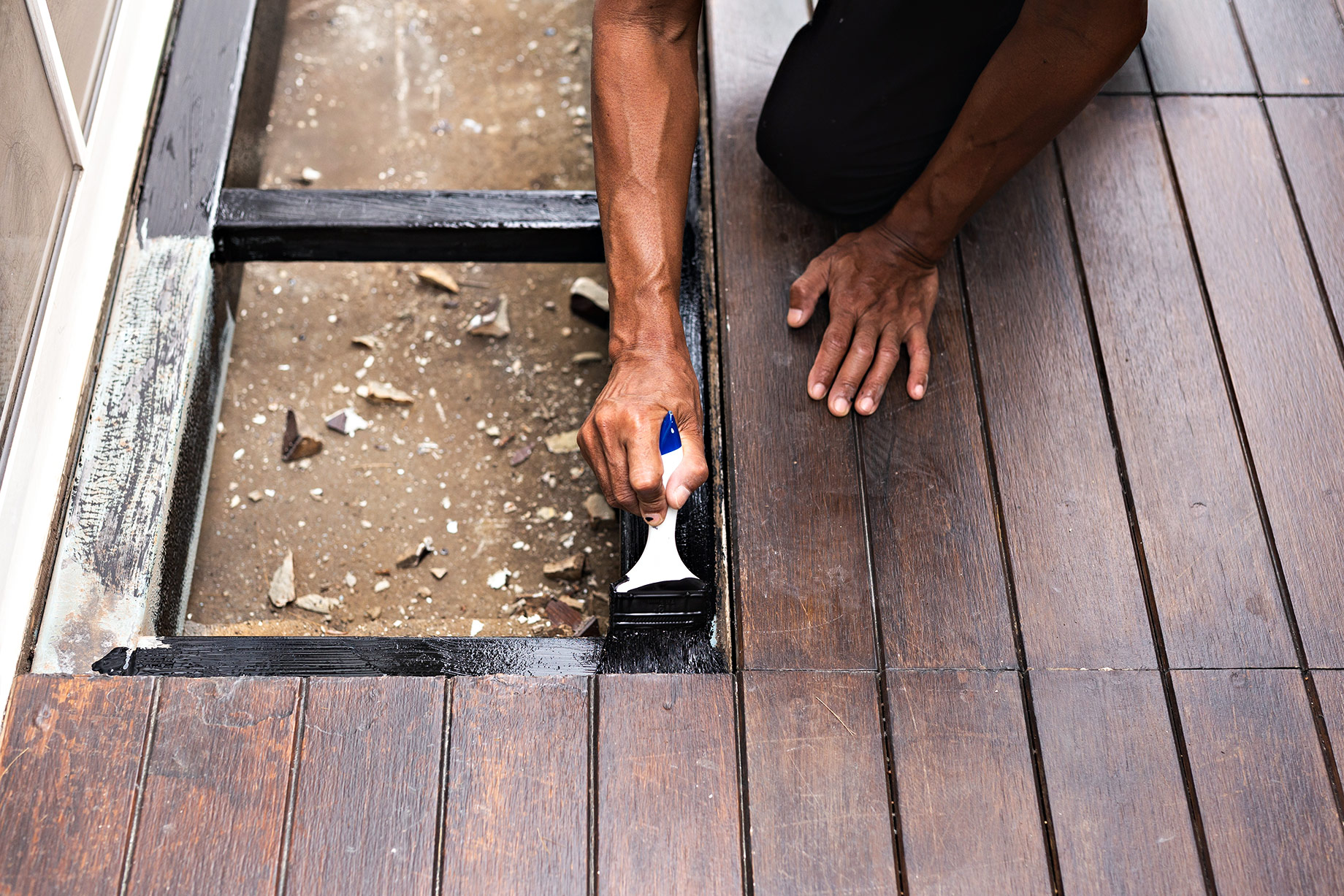 Professional painter applying black anti-rust coating to exposed steel framing beside wooden balcony decking during repair work