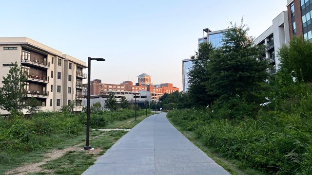 Paved Atlanta Beltline trail between apartment buildings and lush greenery, with path lights and a red-brick tower in the distance.