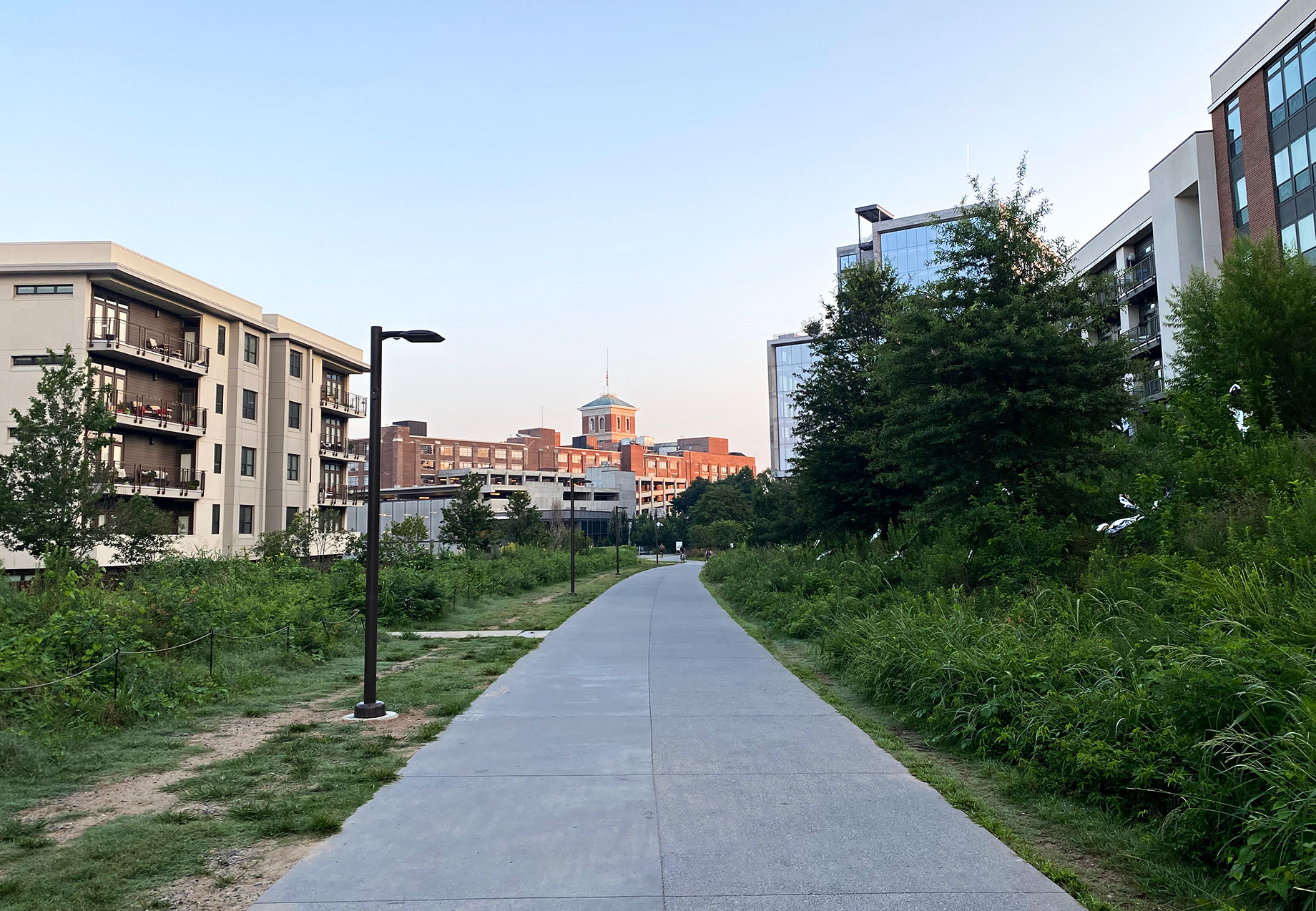 Paved Atlanta Beltline trail between apartment buildings and lush greenery, with path lights and a red-brick tower in the distance.