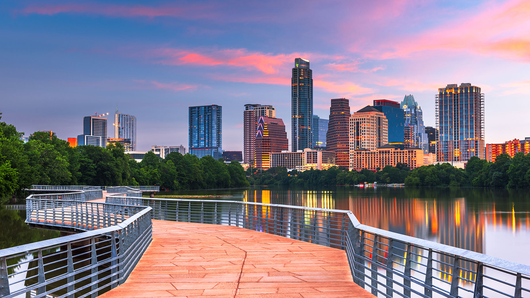 Downtown Austin skyline at sunset reflected across the water, with a waterfront path and city buildings illuminated in the distance.