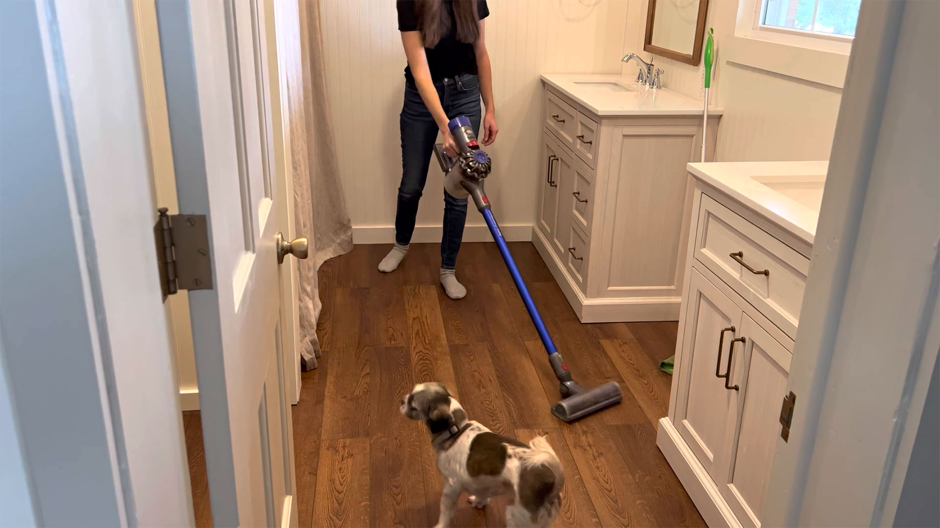 Woman vacuuming a wood-toned bathroom floor beside white vanity cabinets, with a small dog standing nearby.