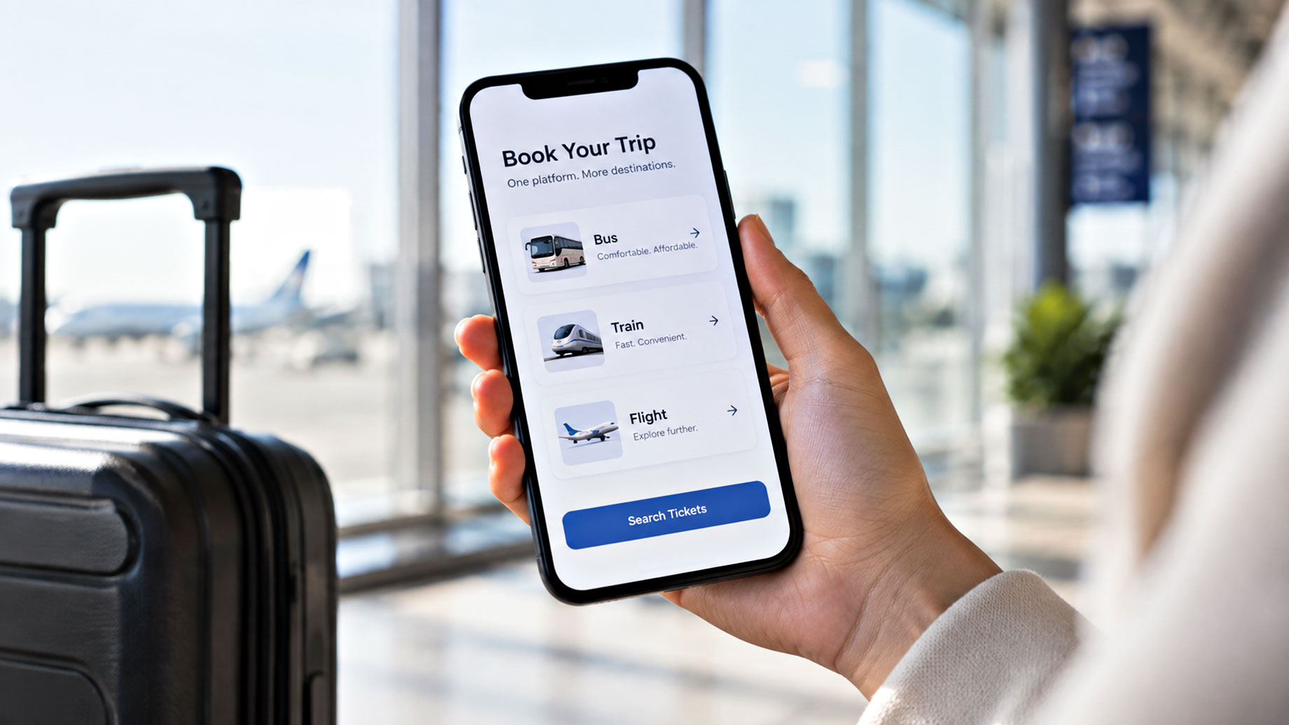 Traveler holding a smartphone with bus, train, and flight booking options beside a suitcase in a bright airport terminal.