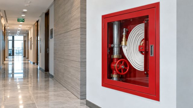 Red fire protection valve cabinet with hose, valve, and piping mounted in a modern commercial building corridor.