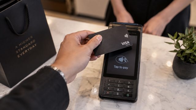 Customer holding a black contactless payment card above a terminal on a stone counter, with a retail assistant in the background.