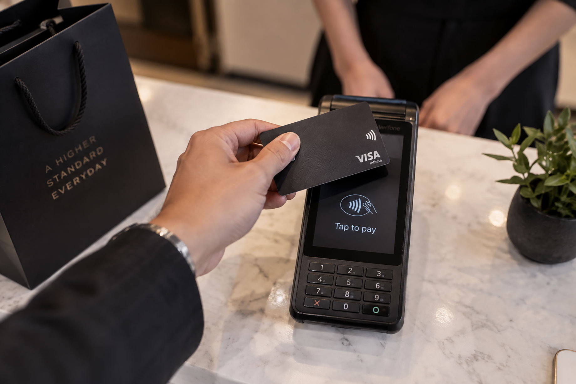 Customer holding a black contactless payment card above a terminal on a stone counter, with a retail assistant in the background.
