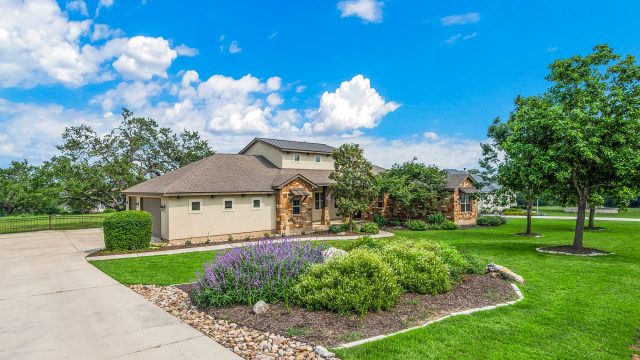 Stone and stucco home in Dallas with a green lawn, flowering garden beds, mature trees, and a curved driveway.