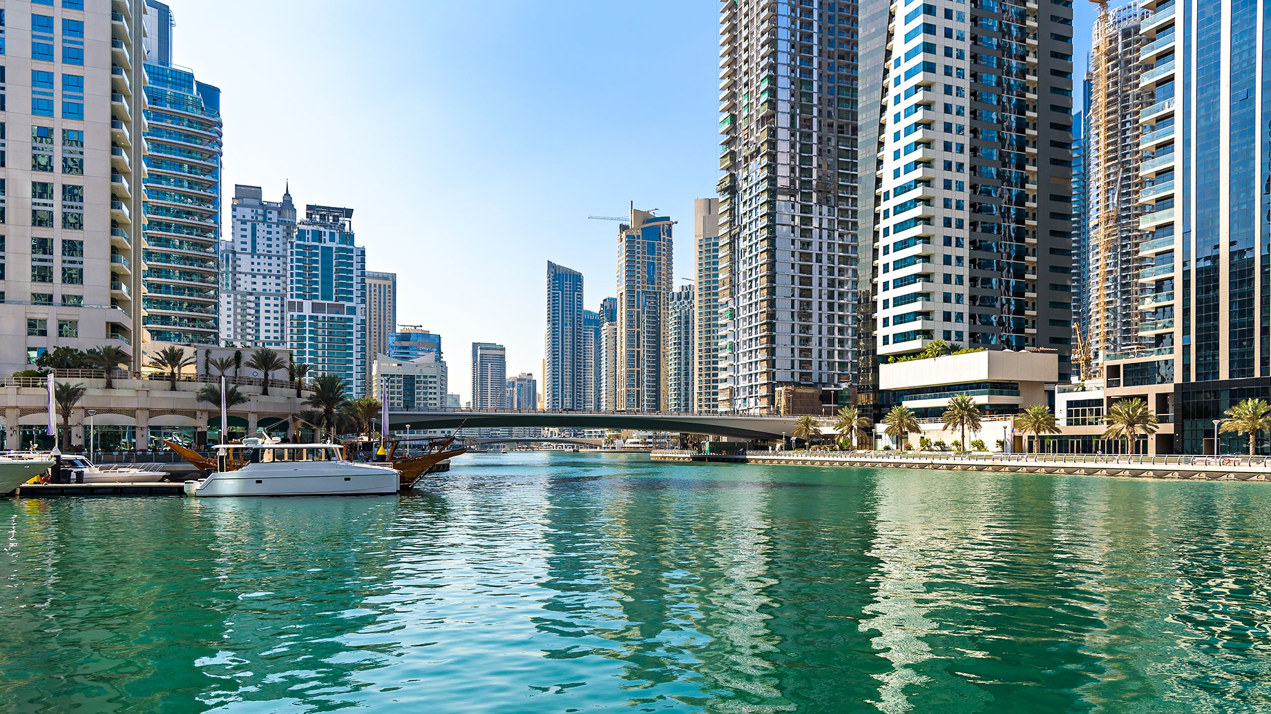 High-rise towers overlooking Dubai Marina’s turquoise canal, with moored boats, palm-lined promenades, and a bridge.