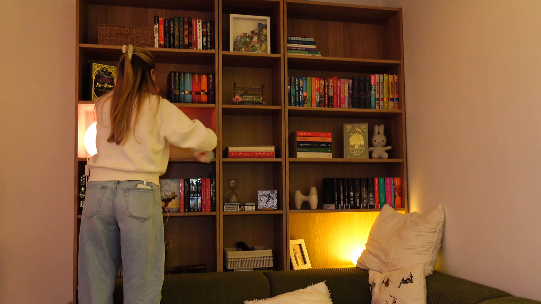 Woman cleaning a wooden bookshelf filled with books and decorative objects behind a green sofa in a warmly lit living room.