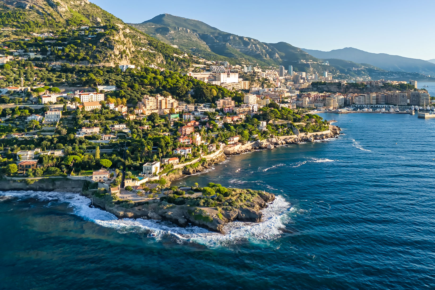 Aerial view of French Riviera villas on green hillsides above rocky headlands and blue Mediterranean water.