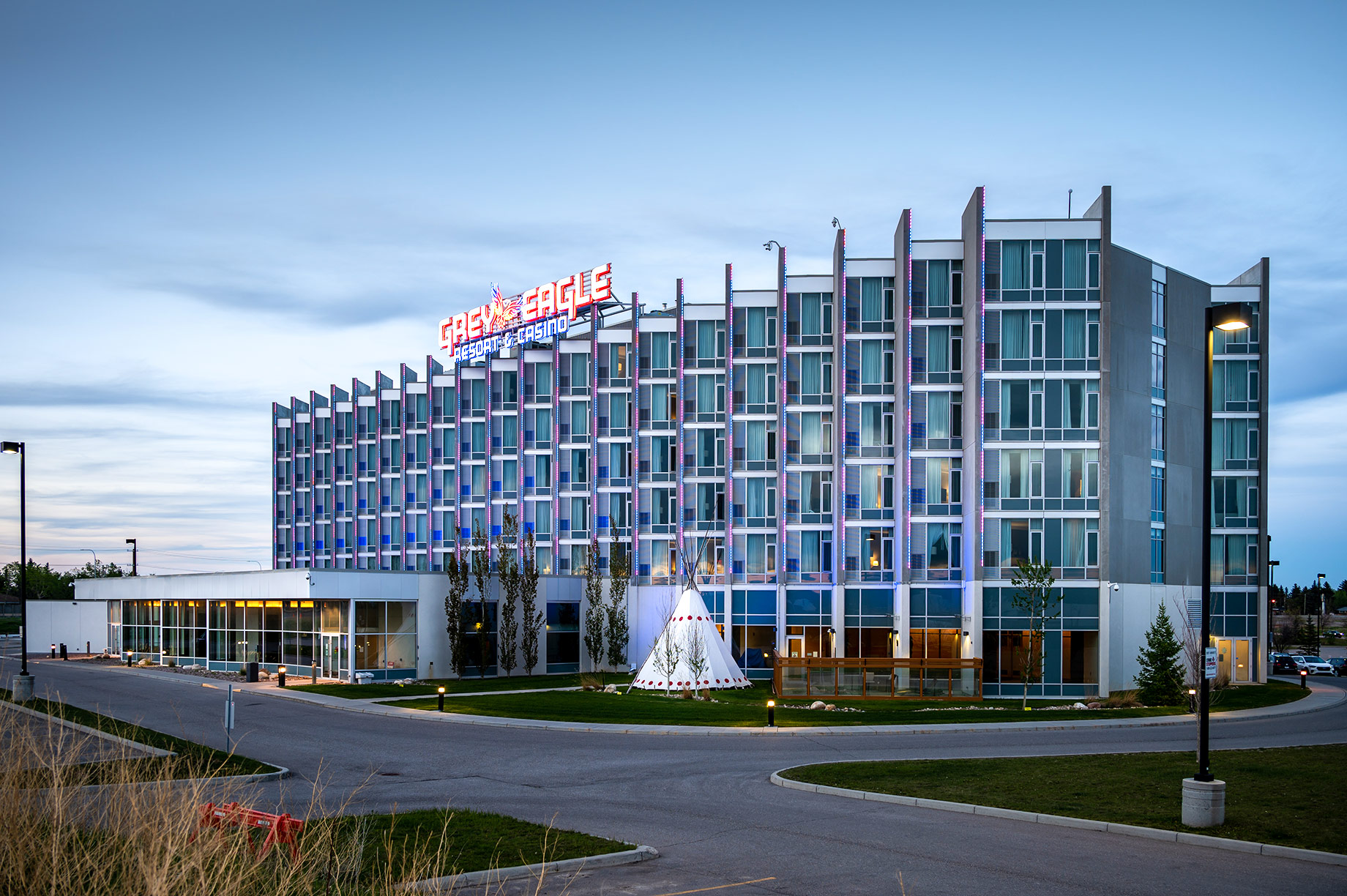 Grey Eagle Resort & Casino hotel exterior illuminated at dusk with colourful vertical lighting and a tipi on the landscaped grounds.
