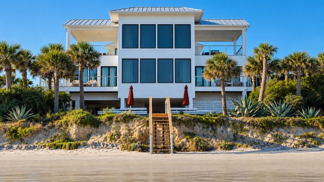 White luxury beachfront home with palm trees, dune landscaping, balconies, and a central staircase leading to the sandy shore.