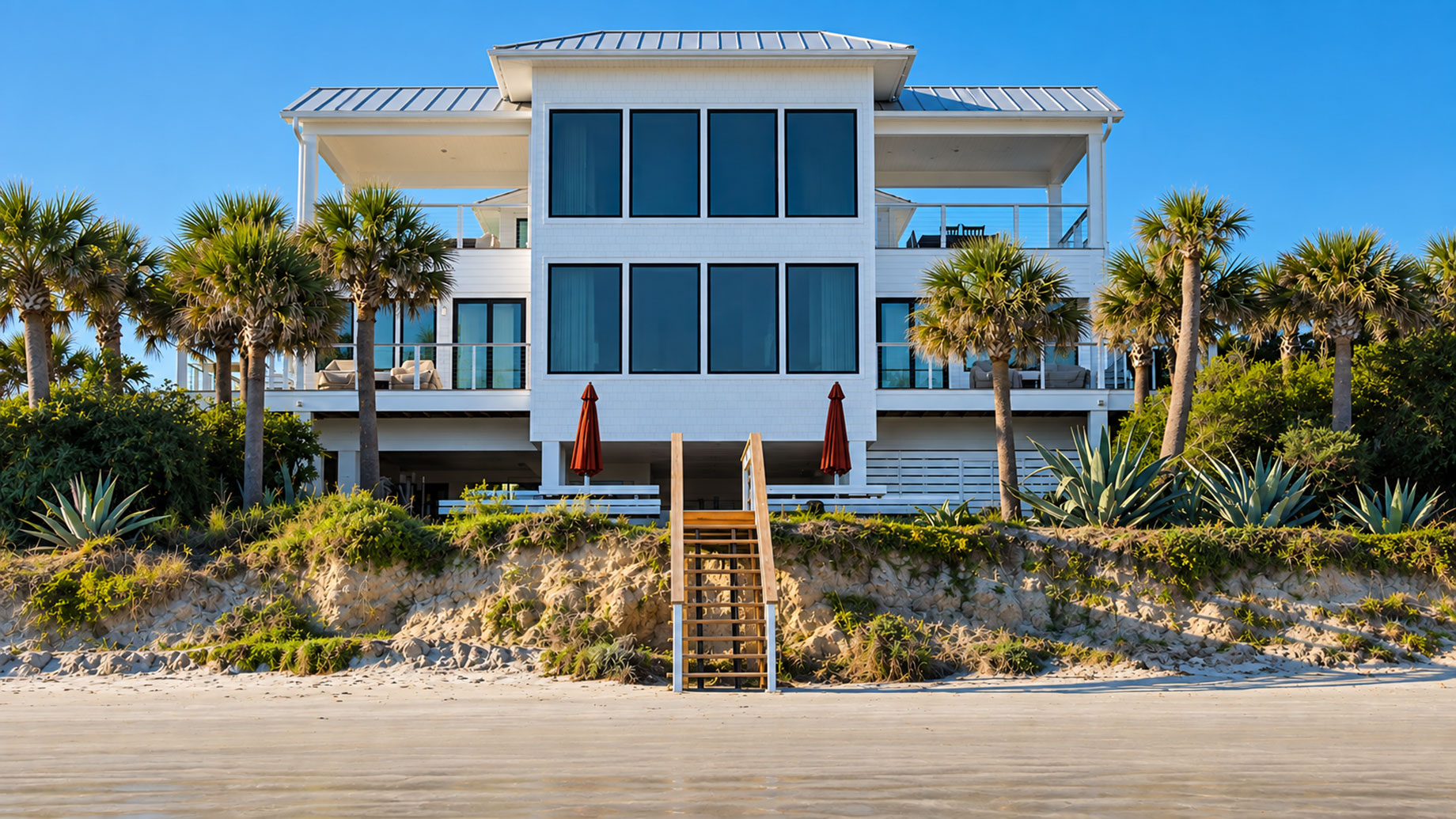 White luxury beachfront home with palm trees, dune landscaping, balconies, and a central staircase leading to the sandy shore.